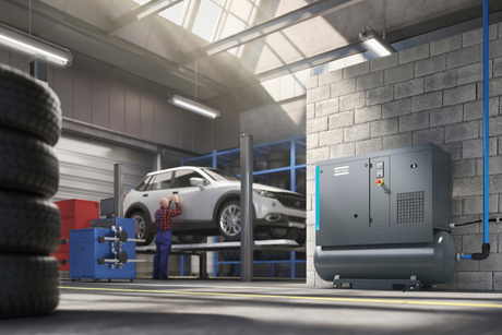 There is a mechanic fixing a white car in the background of the image, the foreground shows a compressor and some tyres. 
