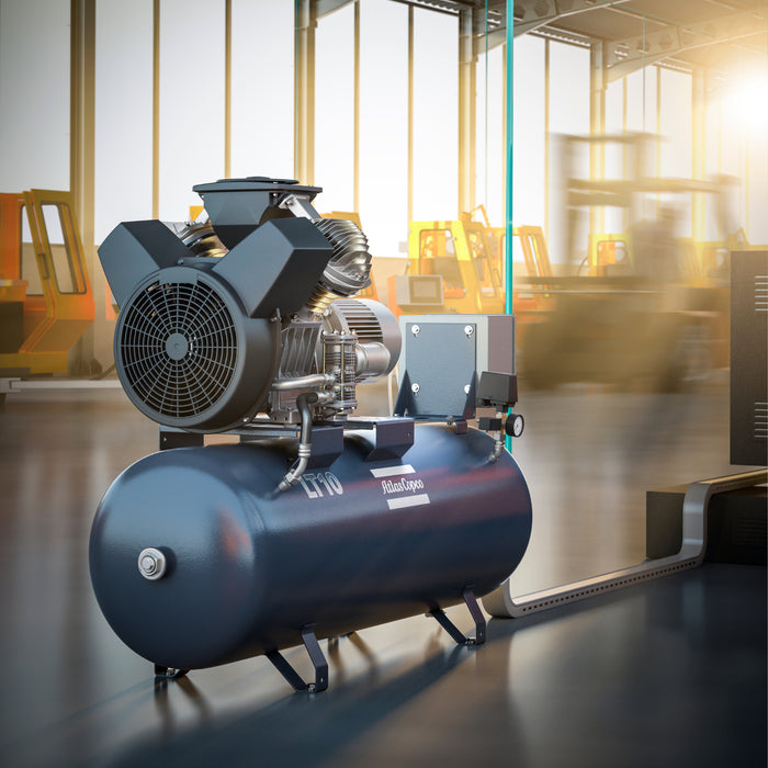Atlas Copco piston compressor on the factory floor attached to a blue air line. The background is blurred with evening sun coming through the window.